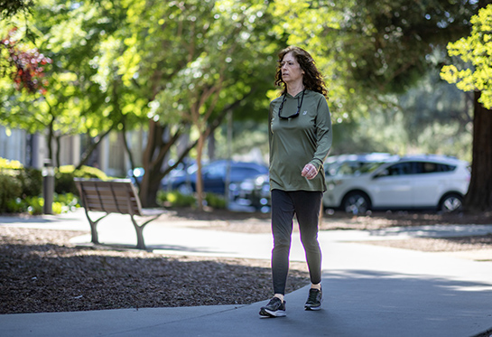 Woman with dark hair, wearing green sweatshirt and navy pants, walks down sidewalk with trees and park bench in background.